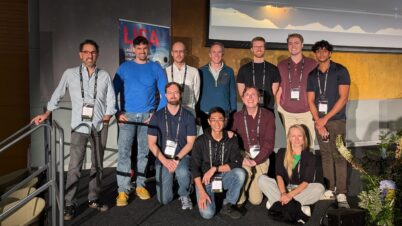 Group photo of the PSSL team at a symposium, with eleven members standing and kneeling in front of a LISA poster. They are wearing conference badges and are in a well-lit room with a projector screen behind them.